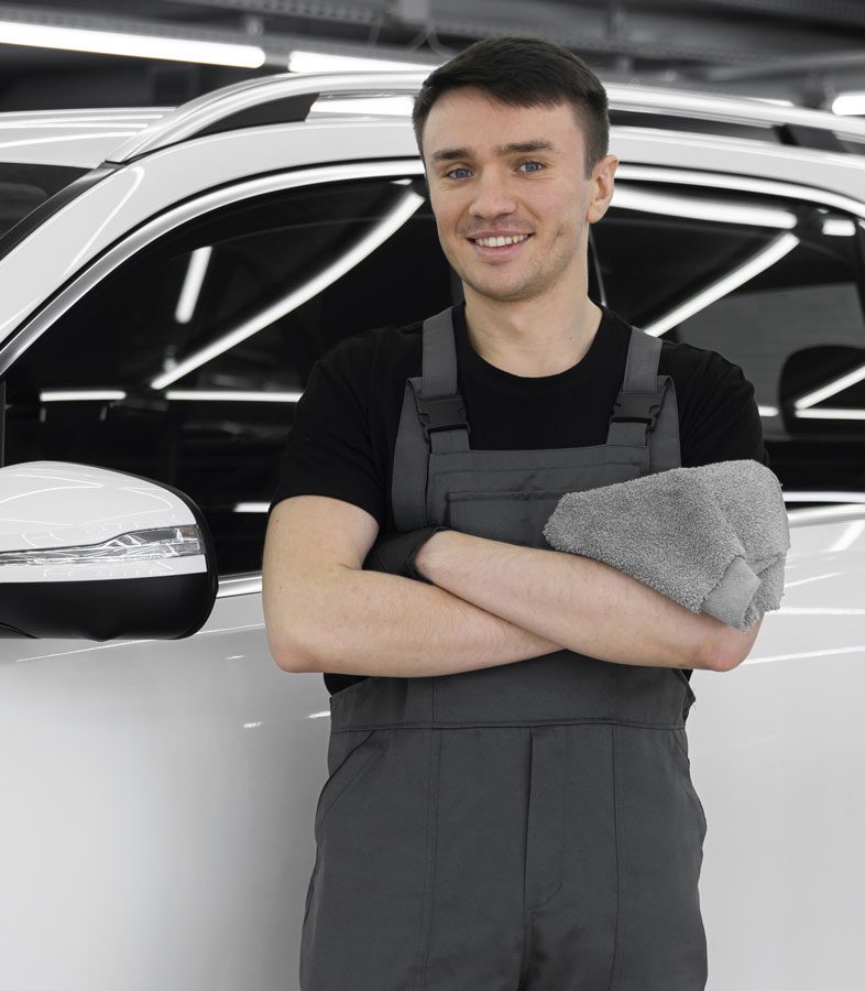 Smiling male car detailer in gray overalls holding a cleaning towel, standing next to a luxury vehicle at Evoke Auto Spa.